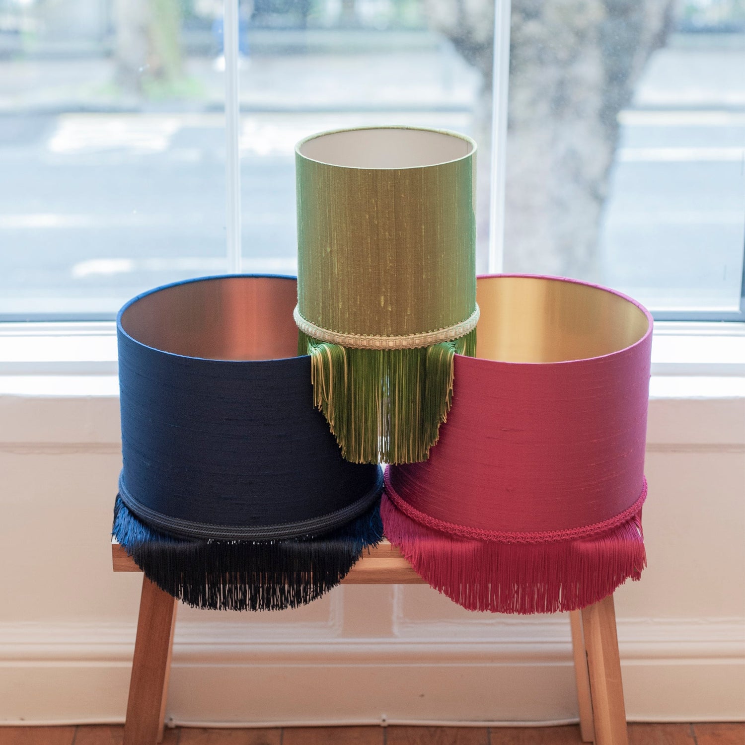 Three colorful lampshades on a wooden stool with a window in the background