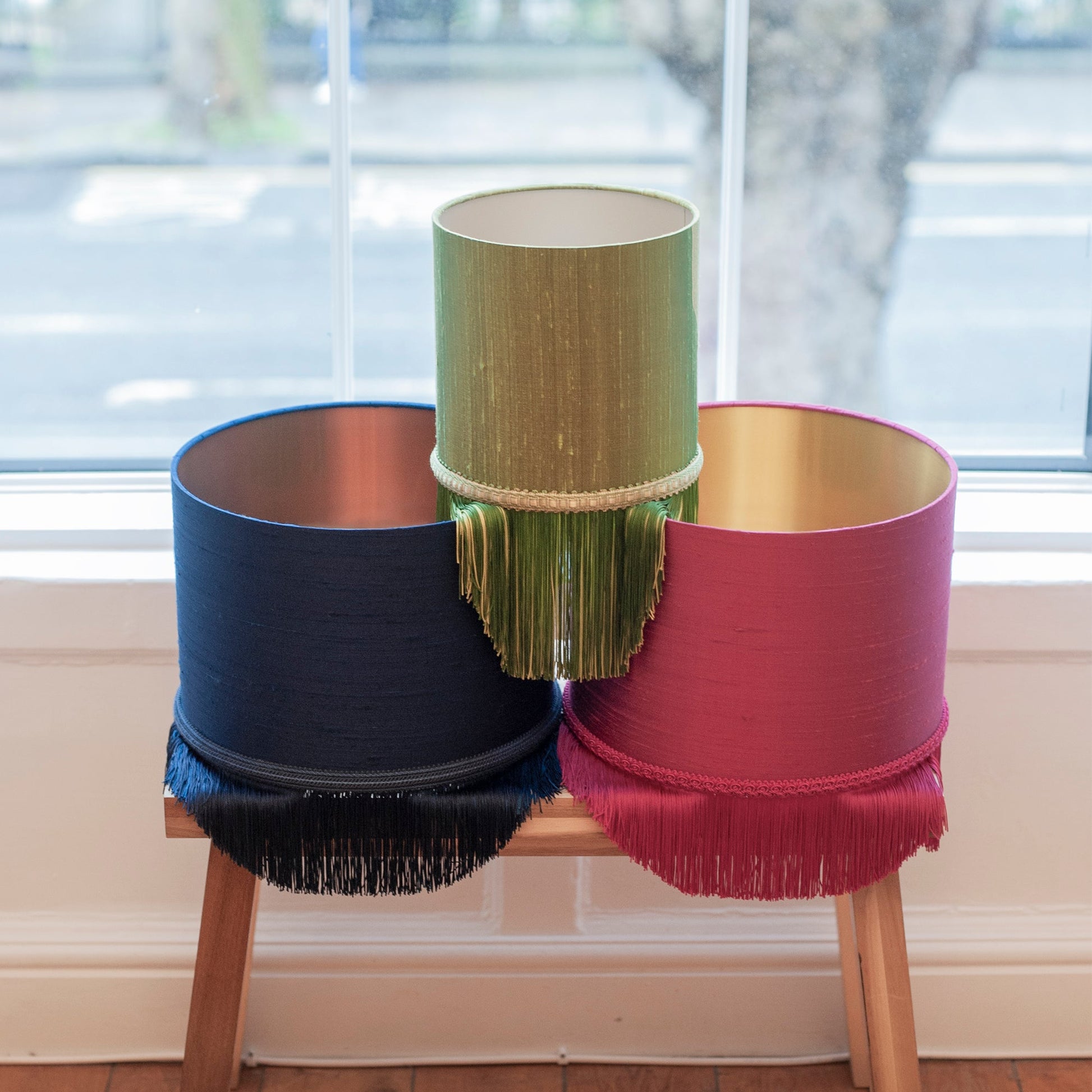 Three colorful lampshades on a wooden stool with a window in the background