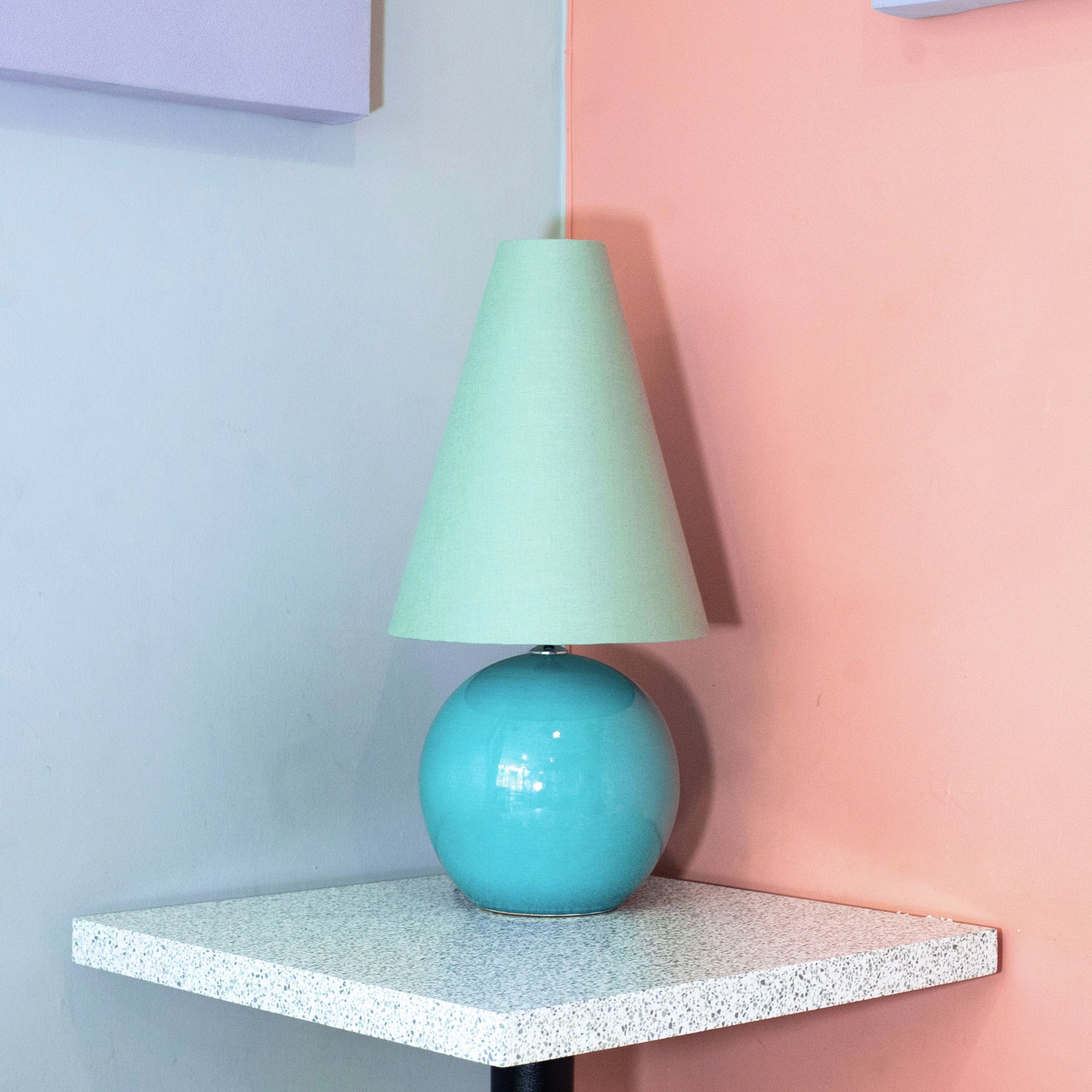 Turquoise lamp with a green shade on a white shelf against a pastel wall.